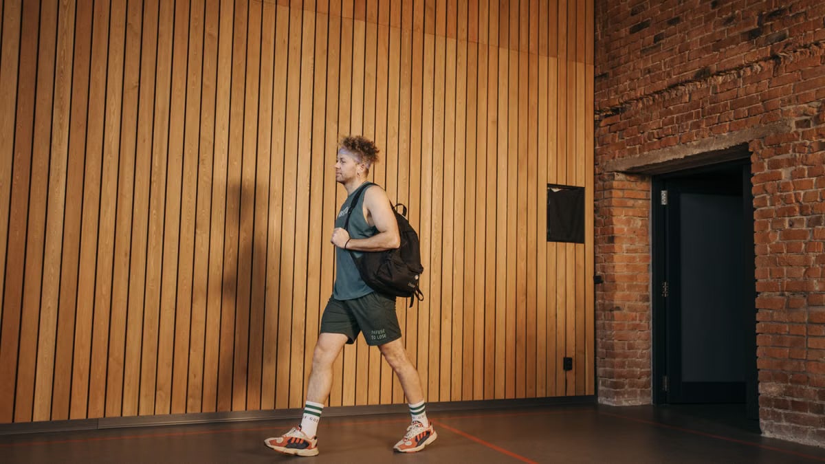Adult man in gym attire walking indoors with a backpack against a wooden wall.